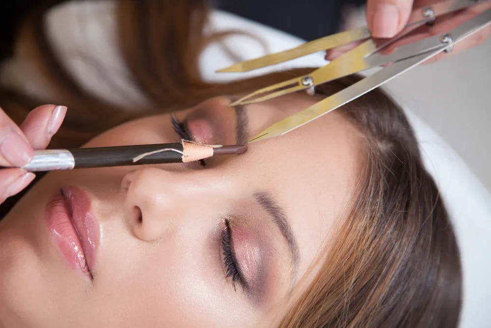 Brow shaping in progress at Envisage Beauty — brow pencil and gold calipers measuring brow arch
