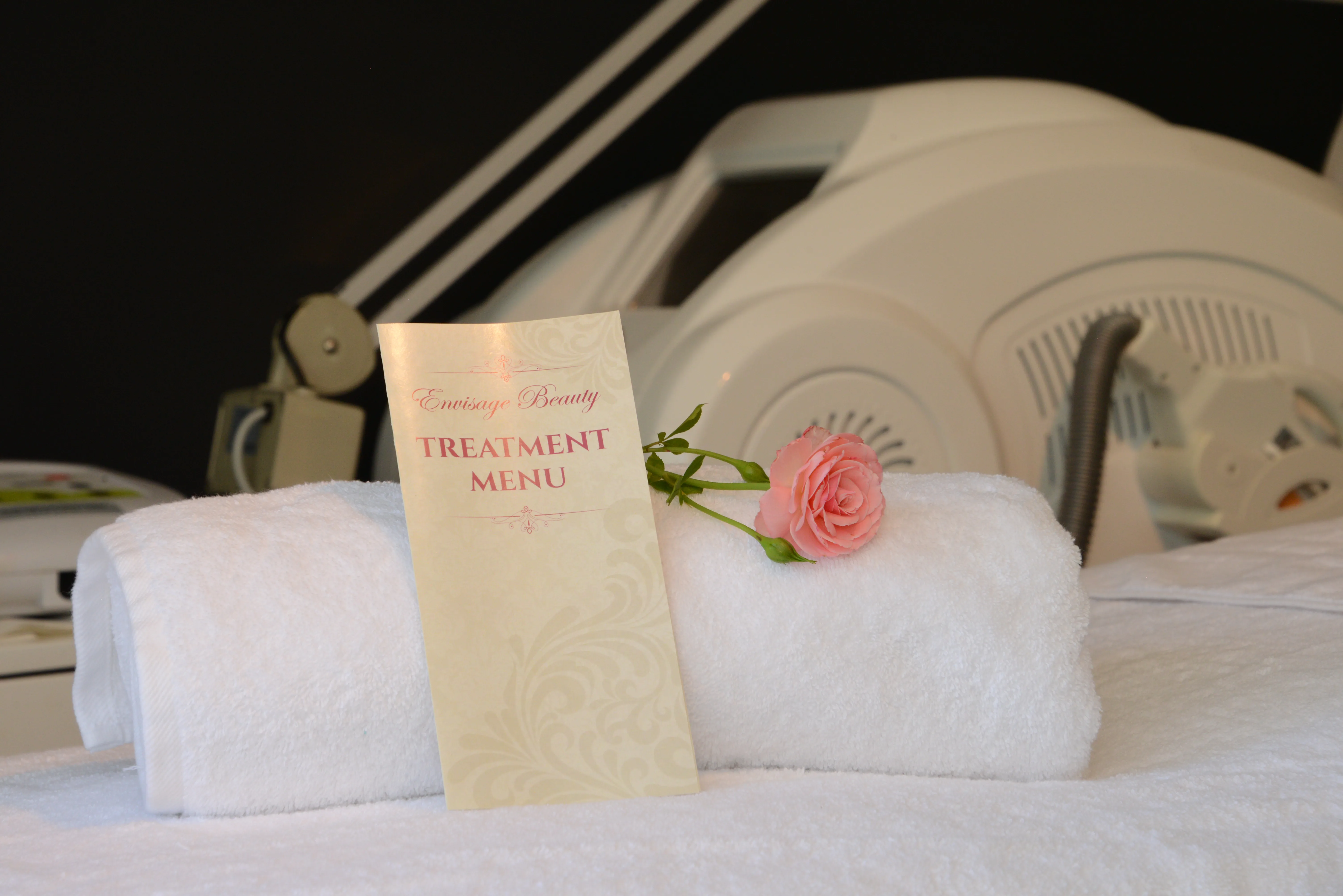 Envisage Beauty treatment menu brochure resting on rolled towels with a rose — representing the salon's full range of in-clinic and retail offerings
