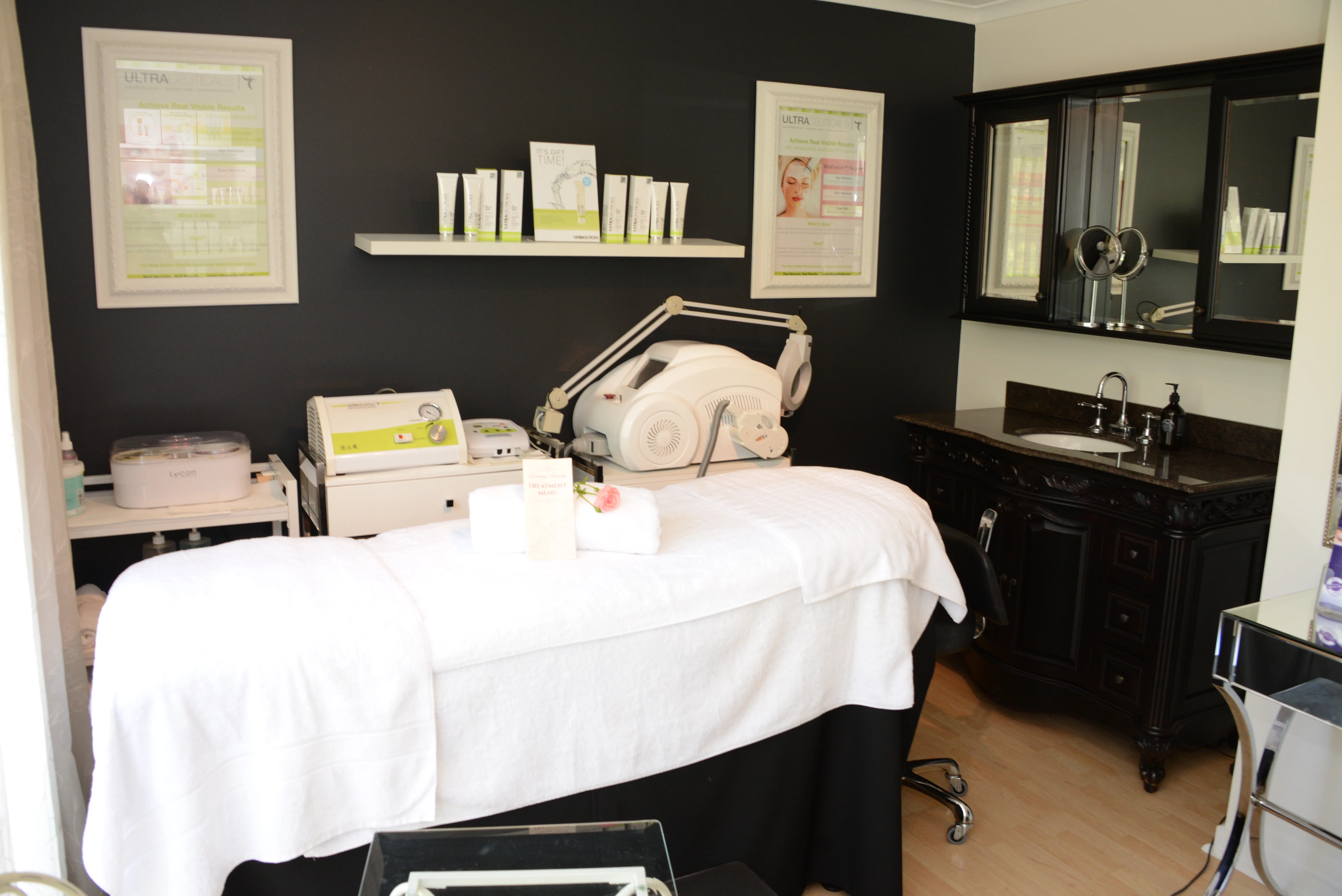Envisage Beauty treatment room — draped bed, treatment machines and framed Ultraceuticals posters on charcoal wall